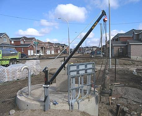 Una grúa pescante eme de radio largo montada en una bóveda de servicios de concreto circular en una obra de fraccionamiento residencial en construcción, con el brazo extendido sobre la abertura de acceso.