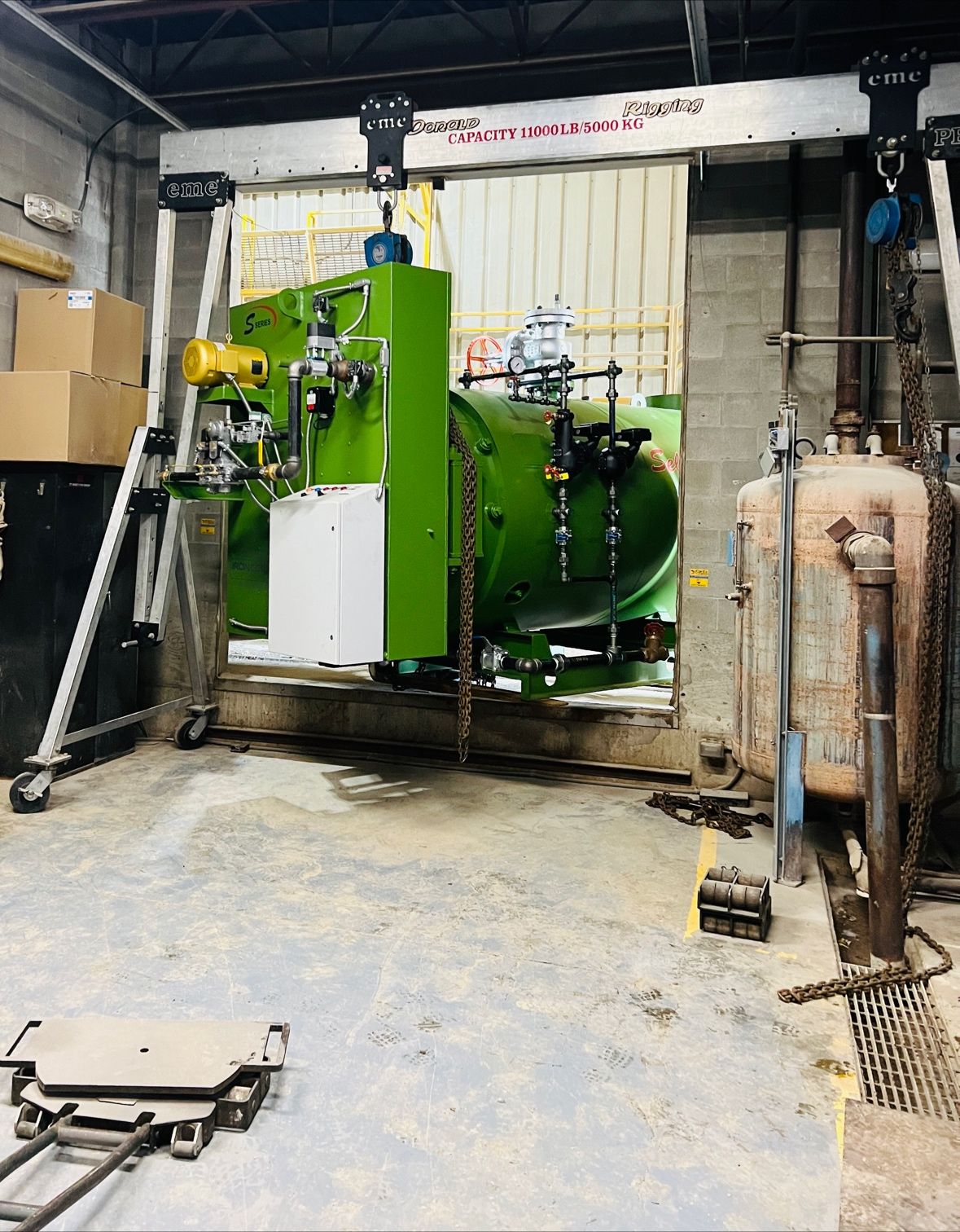 Two eme 11000 lb capacity aluminum gantry cranes positioned around a large green industrial process vessel being lifted into place in a utility / boiler room, with a rusted pressure tank visible to the right.