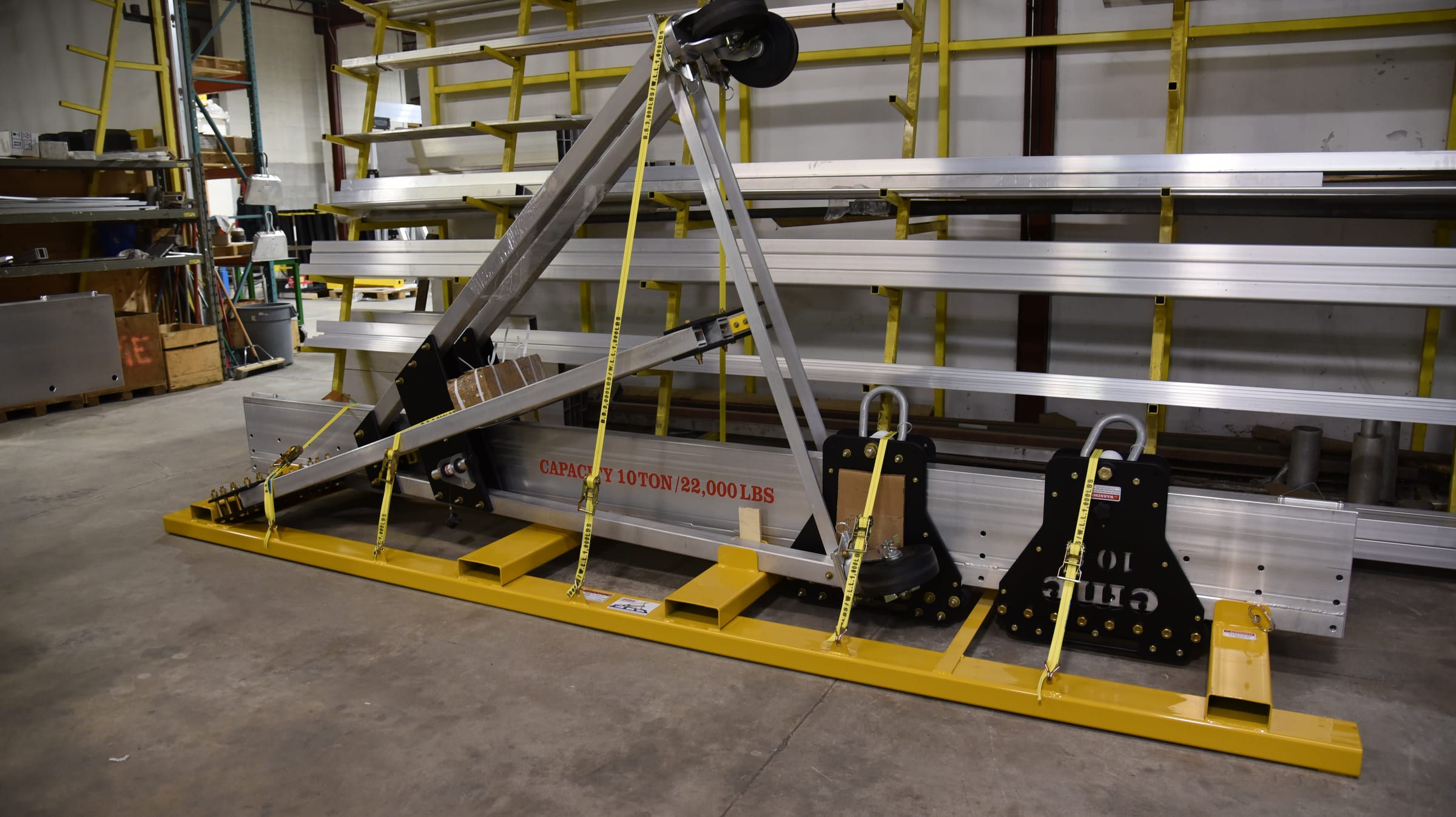 An eme 22000 Series aluminum gantry crane broken down and secured to a yellow steel shipping frame: aluminum beams, leg struts, trolley with CAPACITY 10 TON / 22,000 LBS stamp, and hardware tied down with safety straps for transport.