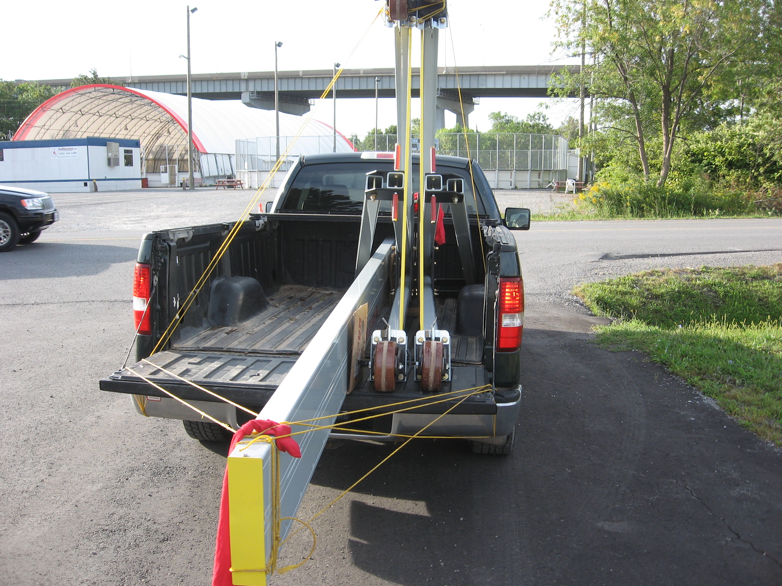 An eme 6600 / 8800 Series gantry crane top beam loaded across a pickup truck bed for site-to-site transport, with the trolley assembly and casters visible at the back of the truck.